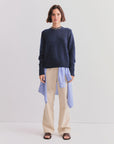 A woman leans against a white wall wearing Kule’s The Cashmere Tatum crewneck in navy, layered over a blue-striped shirt, with beige pants, black sandals, and a simple necklace—an effortless wardrobe staple.