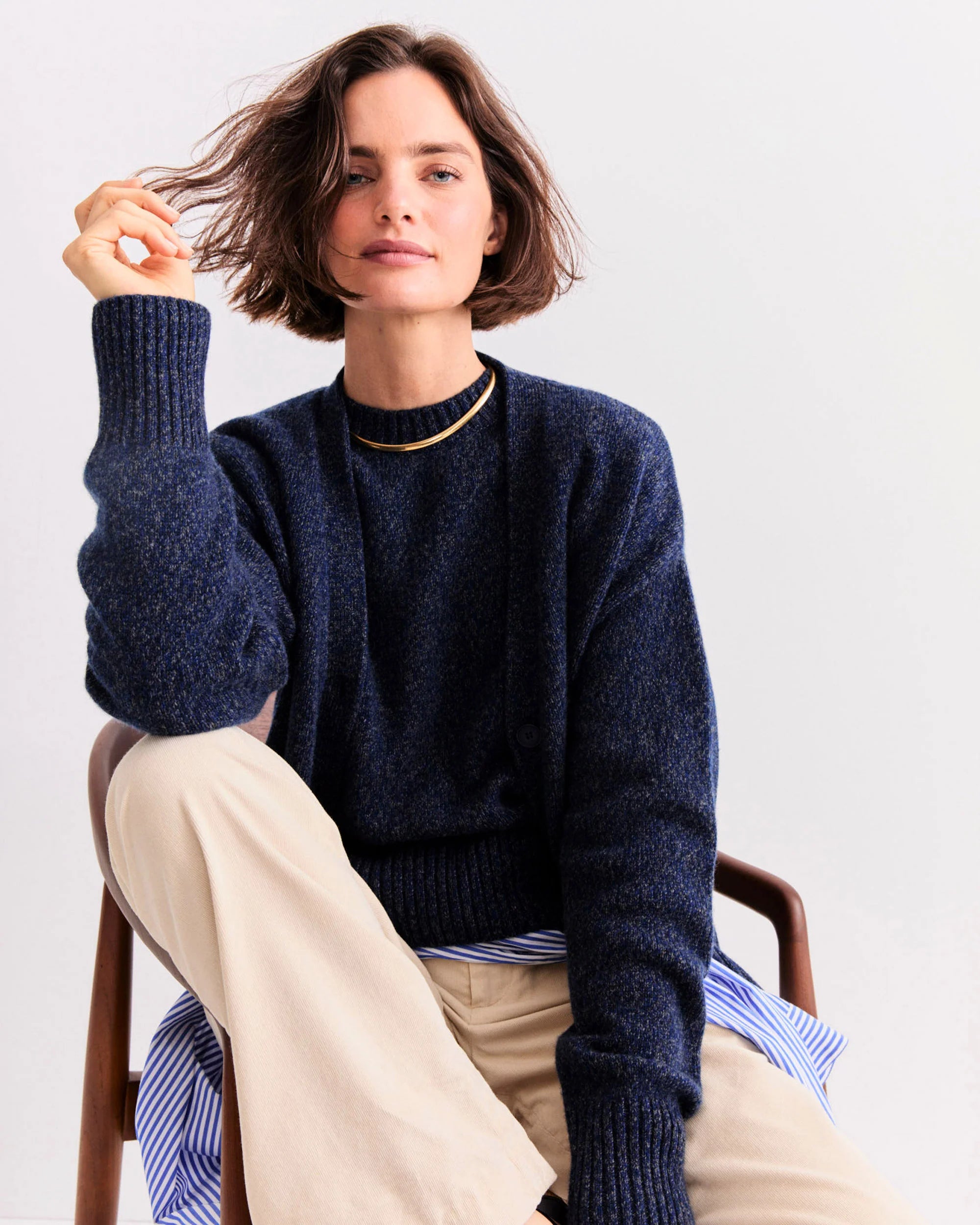 A woman with short brown hair sits on a wooden chair wearing Kule’s The Cashmere Tatum crewneck sweater in navy, paired with beige pants and a gold necklace. She holds her hair, looking confidently at the camera against a plain background.