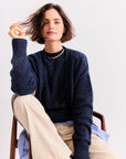 A woman with short brown hair sits on a wooden chair wearing Kule’s The Cashmere Tatum crewneck sweater in navy, paired with beige pants and a gold necklace. She holds her hair, looking confidently at the camera against a plain background.