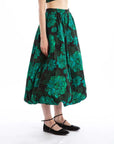 The individual is dressed in a Rhode Cassandra Skirt adorned with the Blossoming Canopy print, showcasing a striking green floral design on a black base. Combined with black ballet flats, their chic ensemble stands out against the simple white background.