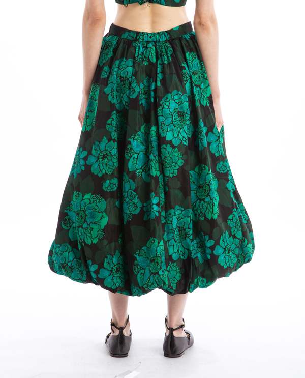 A person is pictured from the back wearing a flowing Rhode Cassandra Skirt featuring the Blossoming Canopy print along with black sandals. The skirt showcases large floral designs and has an elegant bubble hem that falls below the knees, set against a white background.