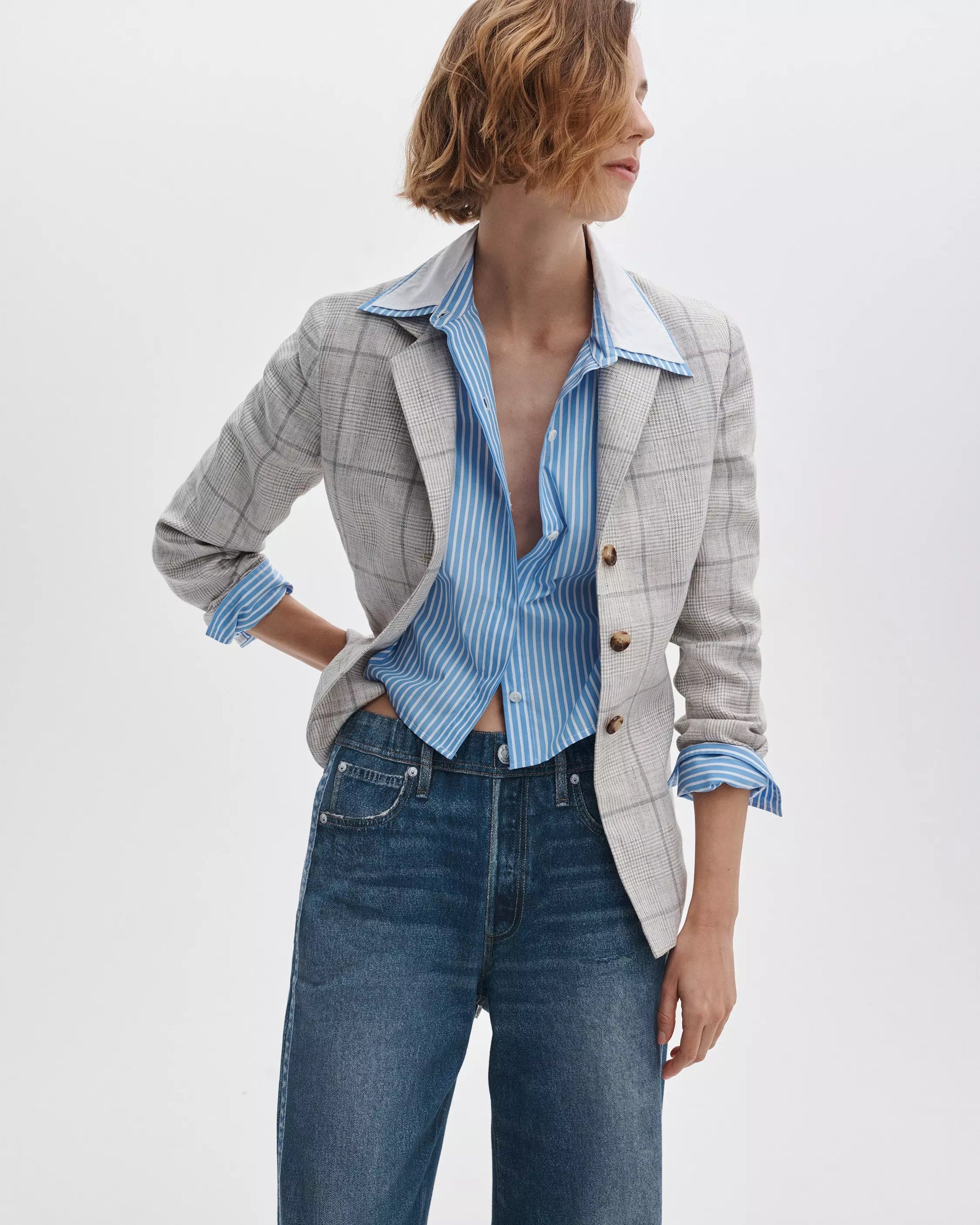 A person wears the Rag & Bone chappell plaid blazer over a blue and white striped shirt with wide lapels, partly unbuttoned, paired with blue jeans, standing against a plain white background.