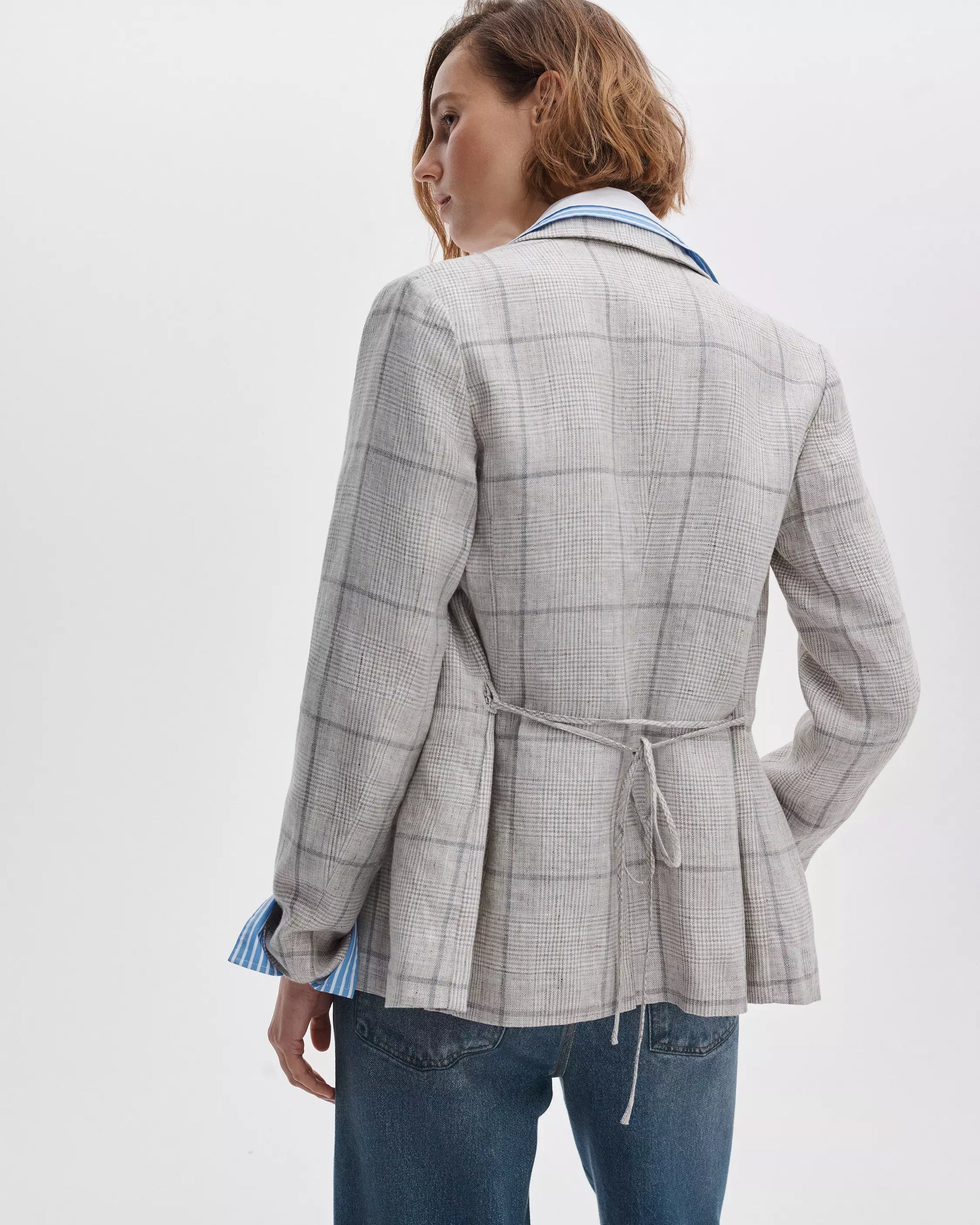 A woman with short, wavy hair is seen from the back wearing the Rag & Bone chappell plaid blazer with a fitted waist tie, a blue collared shirt, and blue jeans against a plain white background.