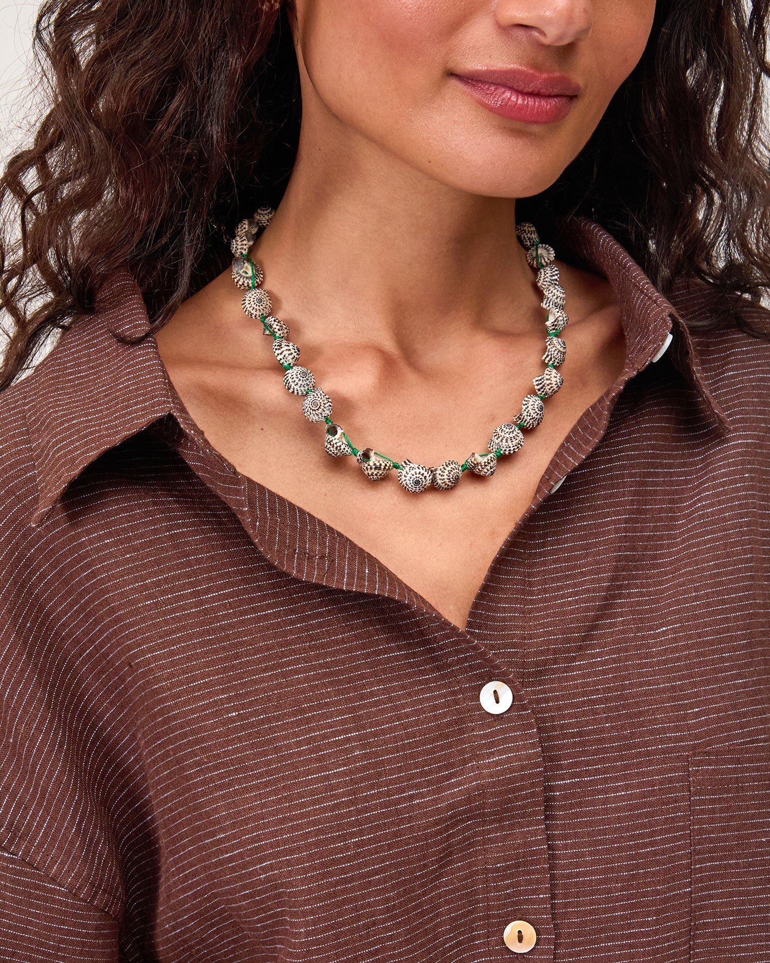 A woman with wavy hair wears a brown pinstriped button-up shirt and the Clare Vivier Checker Shell Necklace. Only her head and upper torso are visible.