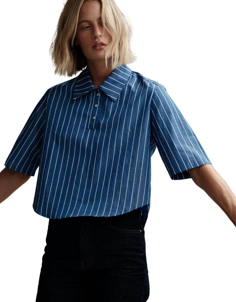 A person wears The Polo Shirt by Cissa, a blue short-sleeved cotton shirt with white vertical stripes, paired with black high-waisted pants against a white background.
