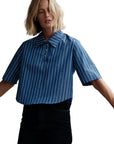 A person wears The Polo Shirt by Cissa, a blue short-sleeved cotton shirt with white vertical stripes, paired with black high-waisted pants against a white background.