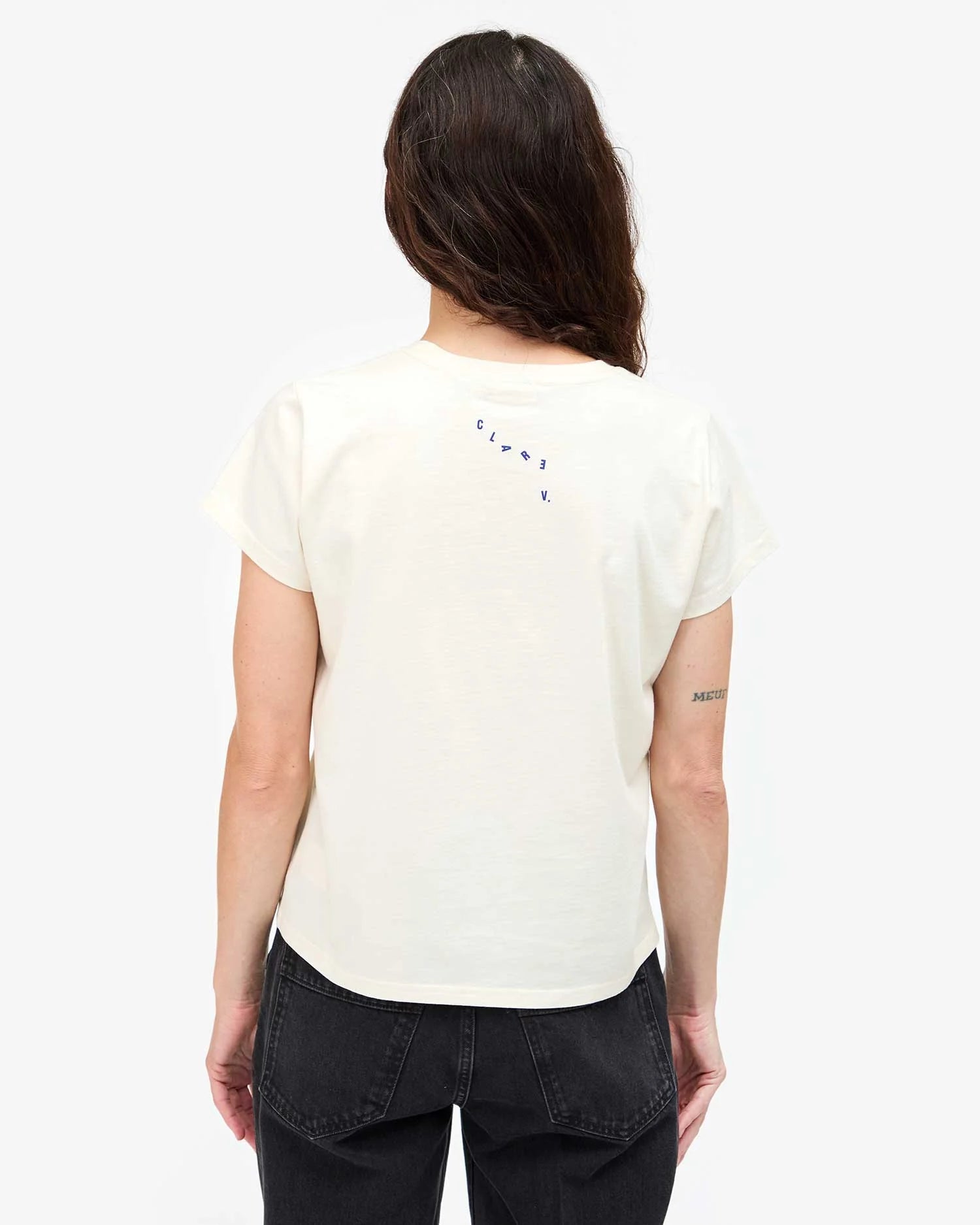 A person with long brown hair faces away, wearing the Clare Vivier Classic Tee CIAO—a lightweight white cotton tee with small blue "CIAO" on the upper back—and black jeans. Their right arm features a small tattoo against a plain white background.