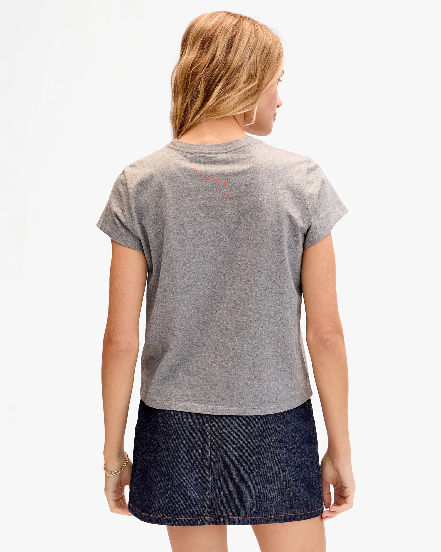 A woman faces away, wearing the Clare Vivier Classic Tee CIAO in gray (100% cotton) with a dark denim skirt. Her blonde hair is loose, she wears a gold bracelet on her right wrist, and the background is plain and light-colored.