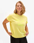 A person with shoulder-length hair stands confidently in Clare Vivier's Classic Tee, a bright yellow 100% cotton T-shirt featuring "DÉJÀ VU" printed in lighter yellow on the front. Paired with a black skirt, they present a stylish ensemble against a plain white background.