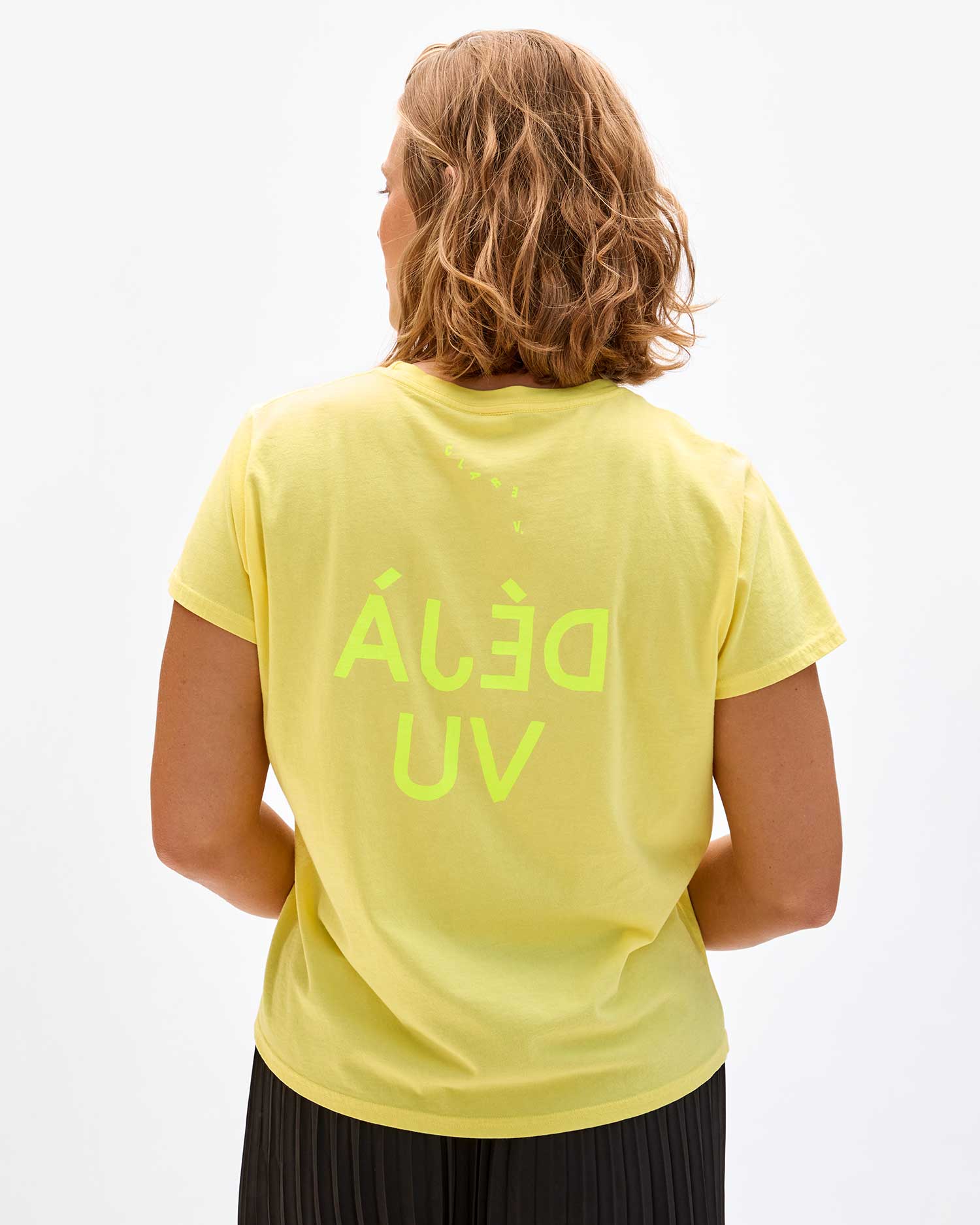 A person with shoulder-length hair is seen from the back, wearing a light yellow Classic Tee by Clare Vivier made of 100% cotton with neon green text "ÁLÉĐ UV" printed in reverse. The t-shirt is paired with dark pants, creating a sense of déjà vu as if this casual look has been encountered before against the plain white background.