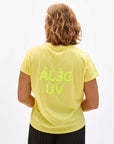 A person with shoulder-length hair is seen from the back, wearing a light yellow Classic Tee by Clare Vivier made of 100% cotton with neon green text "ÁLÉĐ UV" printed in reverse. The t-shirt is paired with dark pants, creating a sense of déjà vu as if this casual look has been encountered before against the plain white background.