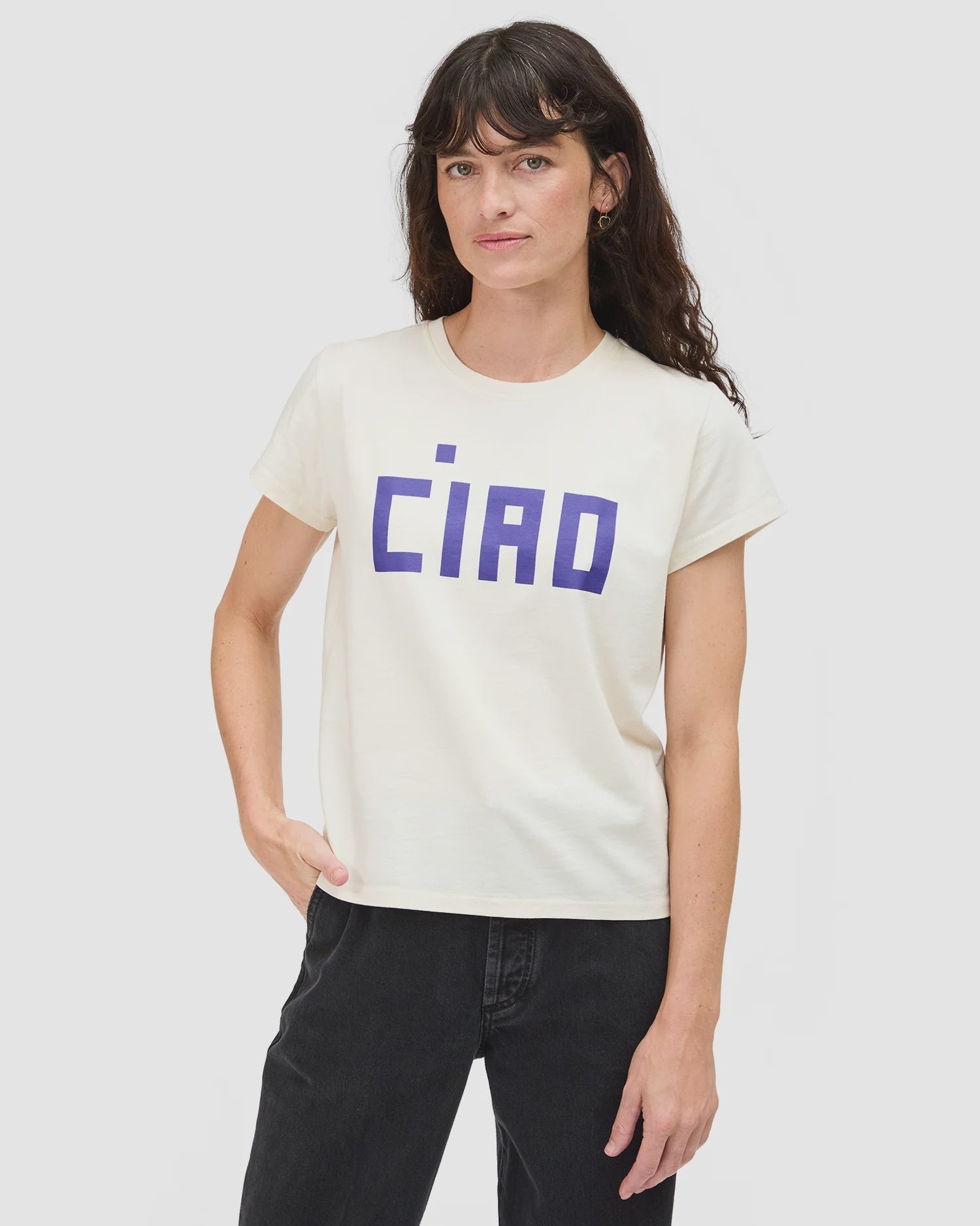 A person with long brown hair wears the Clare Vivier Classic Tee CIAO, a white short-sleeve lightweight cotton T-shirt with bold blue "CIAO" lettering, paired with black pants and standing against a plain light gray background.