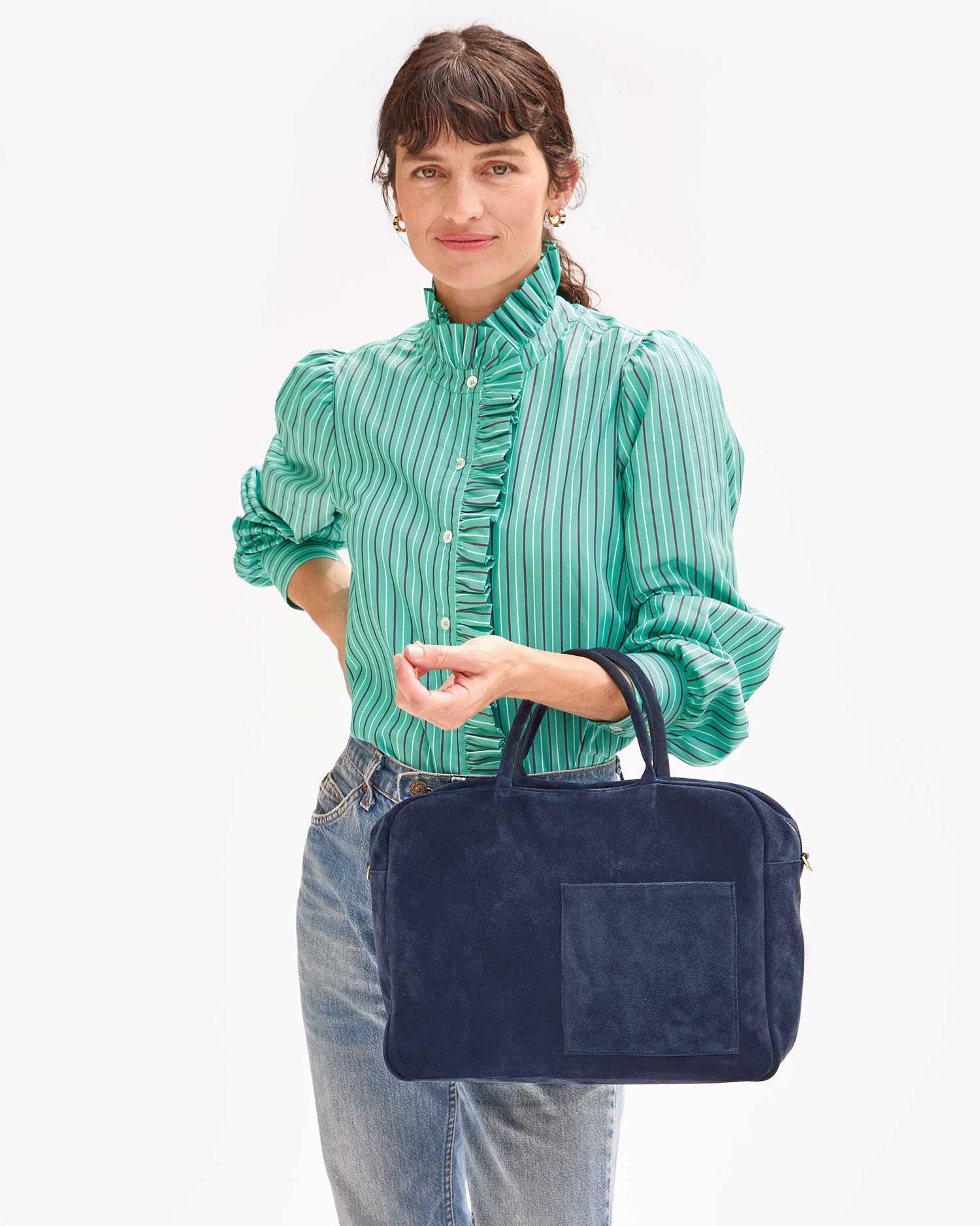 A person stands against a light background wearing a green, striped blouse with ruffled details and blue jeans, holding the Claude Italian suede bag from Clare Vivier. The individual has a neutral expression and hair pulled back with bangs.