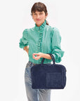 A person stands against a light background wearing a green, striped blouse with ruffled details and blue jeans, holding the Claude Italian suede bag from Clare Vivier. The individual has a neutral expression and hair pulled back with bangs.