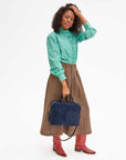 A person poses stylishly against a white background, dressed in a green striped blouse, brown skirt, and red boots. They hold the elegant Claude bag by Clare Vivier in one hand while the other rests on their curly hair.