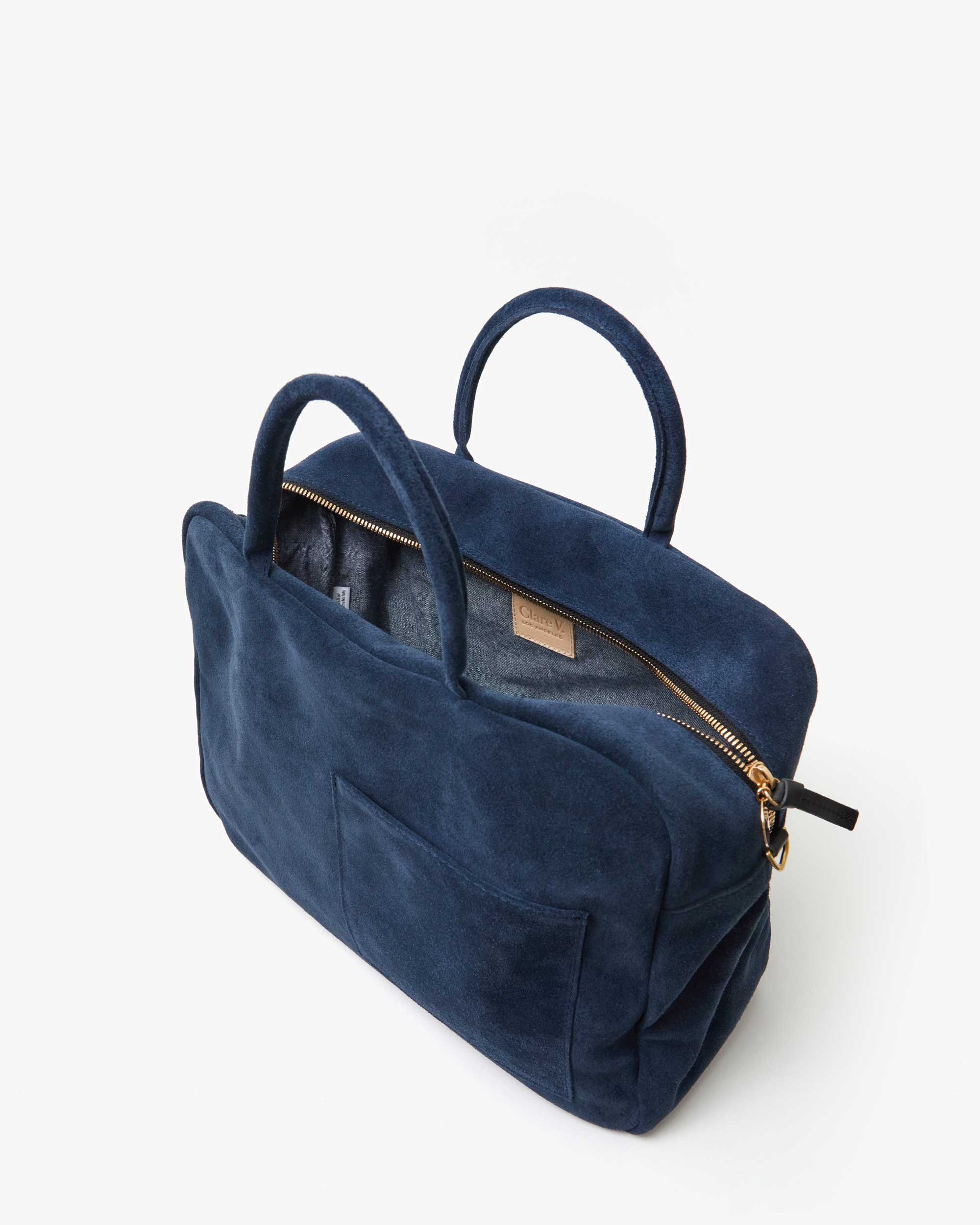 The Claude by Clare Vivier is a navy blue Italian suede handbag with two handles and a gold zipper. It features a front pocket and a partially open top, revealing a gray interior. Perfect for chic outings, this versatile bag is set against a plain white background.