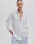 A woman poses against a plain background wearing the Rag & Bone connie shirt in white cotton with an open collar, paired with light blue and white striped pants, hands in pockets and a neutral expression.