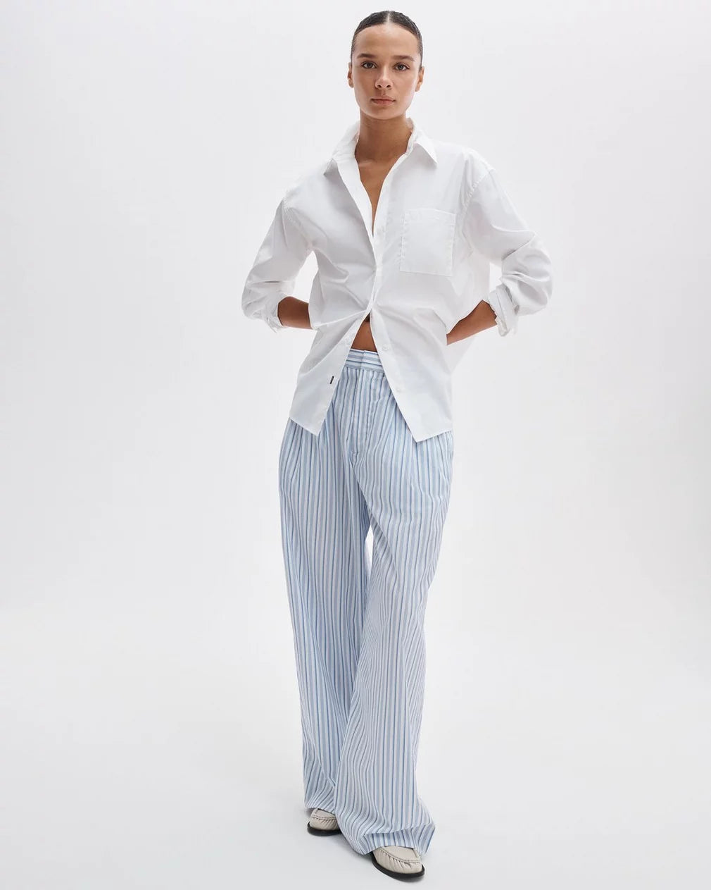 A person stands against a plain white background wearing the Rag &amp; Bone connie shirt, a relaxed-fit cotton style with rolled sleeves, paired with light blue and white striped wide-leg pants and white shoes.