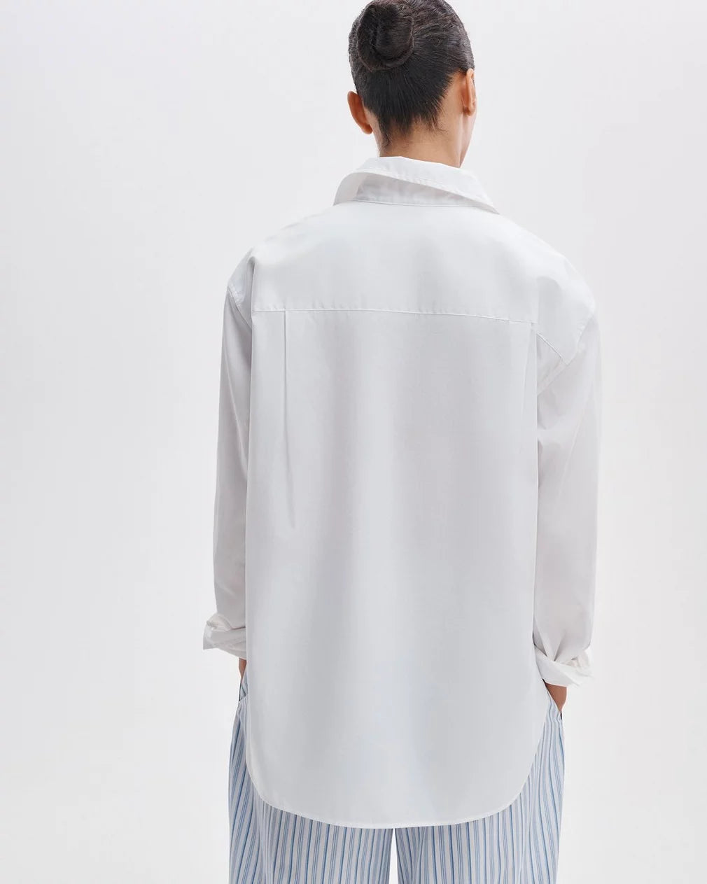 A person with dark hair in a bun, seen from behind, wears the Rag &amp; Bone connie shirt and light blue and white striped pants while standing against a plain light background.