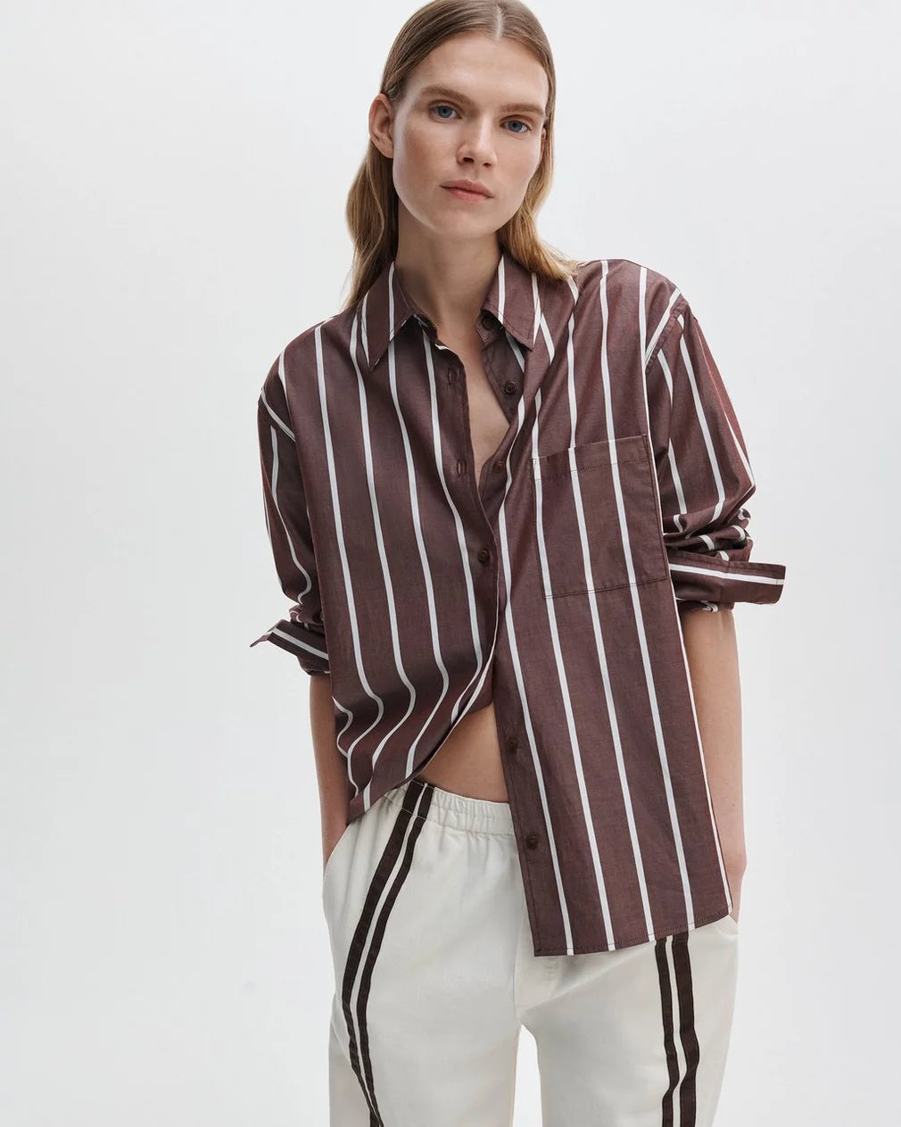 A woman with straight, light brown hair models the Rag & Bone Connie striped shirt, a relaxed-fit cotton poplin top, paired with white pants featuring dark stripes, standing with one hand in her pocket against a plain white background.