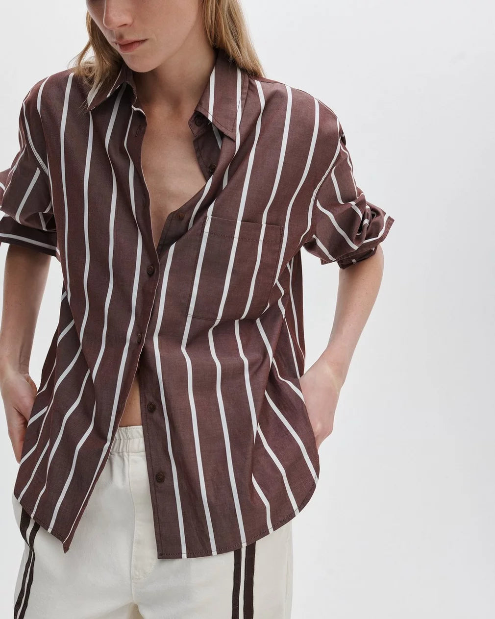 A person wears the Rag & Bone connie striped shirt, a relaxed fit cotton poplin style with white vertical stripes, and off-white pants with a brown side stripe. The shirt is partially unbuttoned with rolled-up sleeves, set against a plain white background.