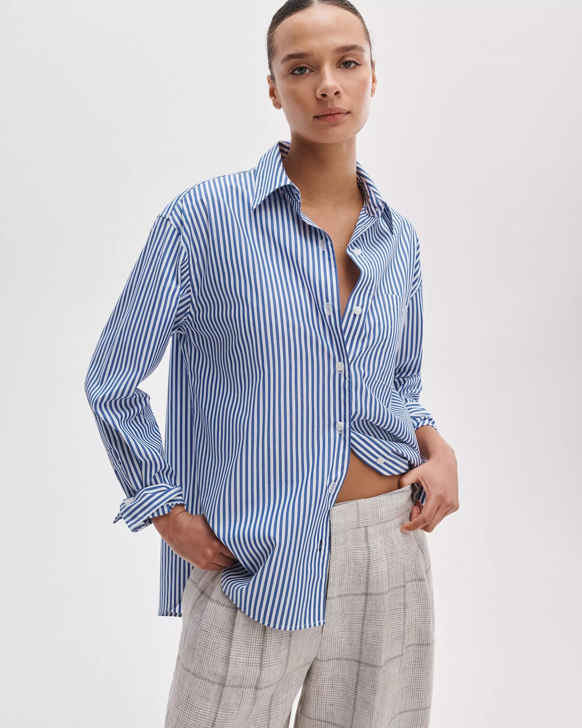 Wearing the Rag & Bone connie striped shirt in blue and white cotton poplin, a person poses with one hand in their pocket against a plain white background, paired with light plaid trousers.