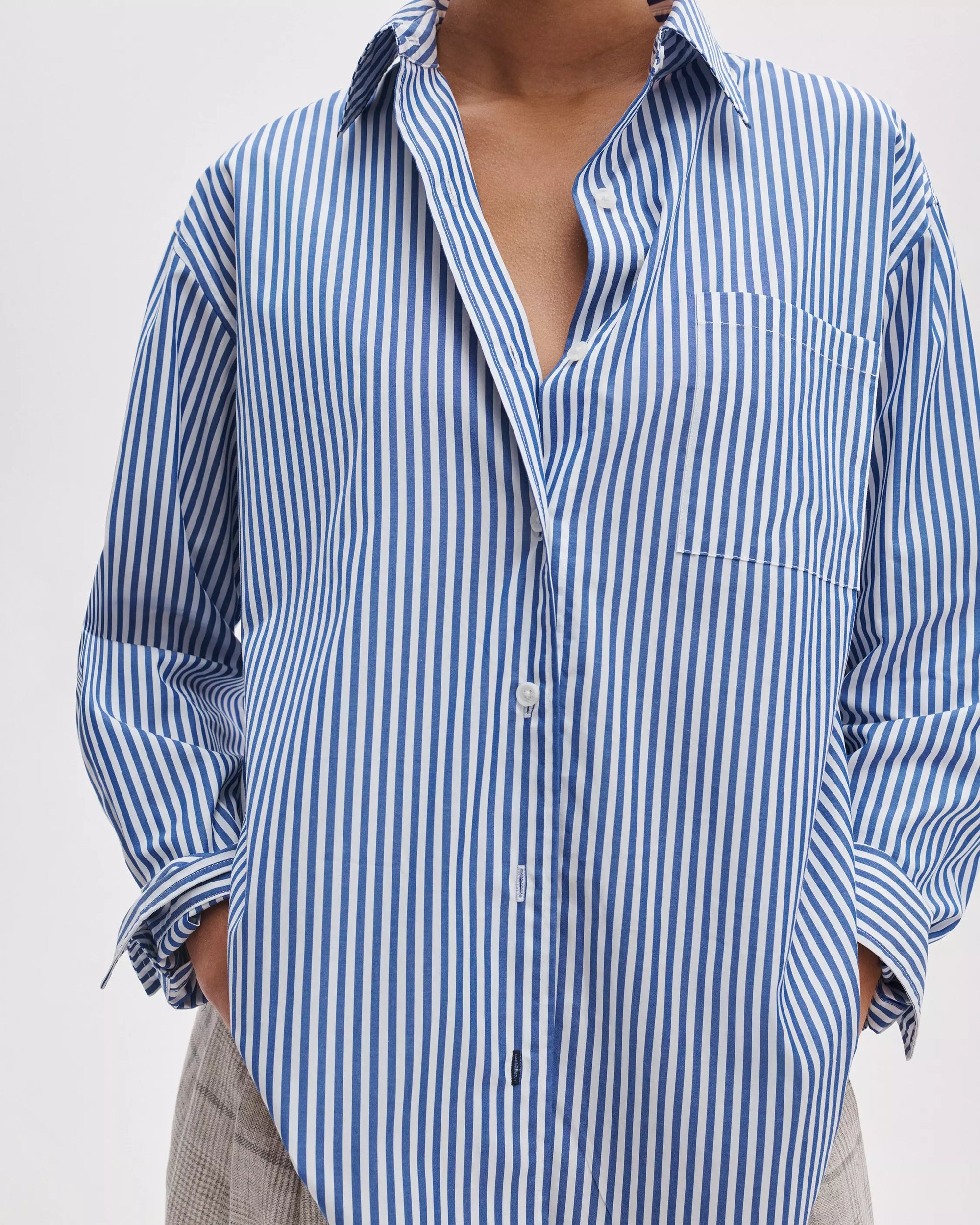 A person wears the Rag & Bone connie striped shirt, a relaxed fit blue and white cotton poplin with vertical stripes, top buttons undone and hands in pants pockets. The image is cropped at the shoulders and thighs.