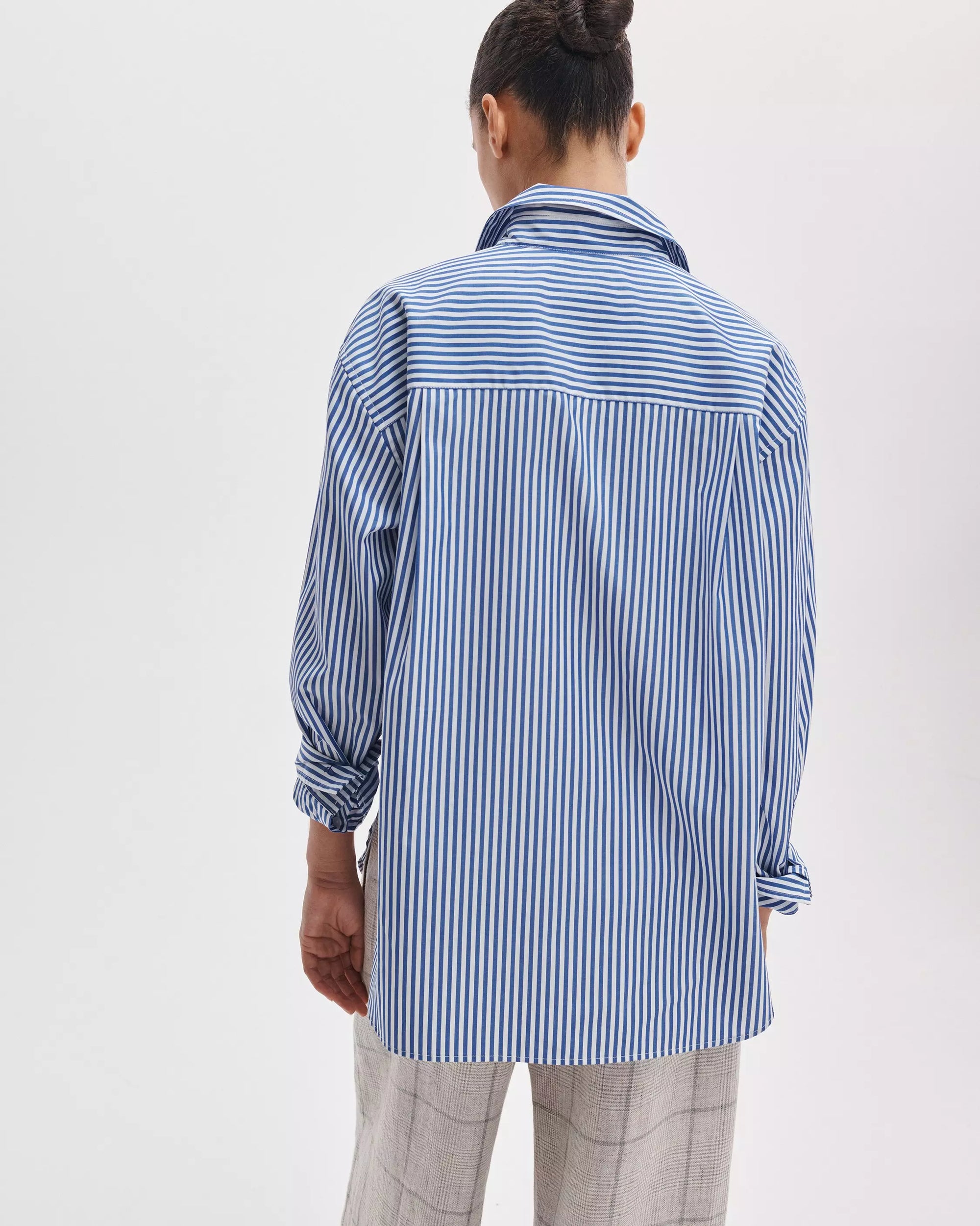 A person faces away, wearing the Rag & Bone connie striped shirt—a relaxed-fit blue and white cotton poplin button-up—paired with light gray plaid pants, set against a plain white background.