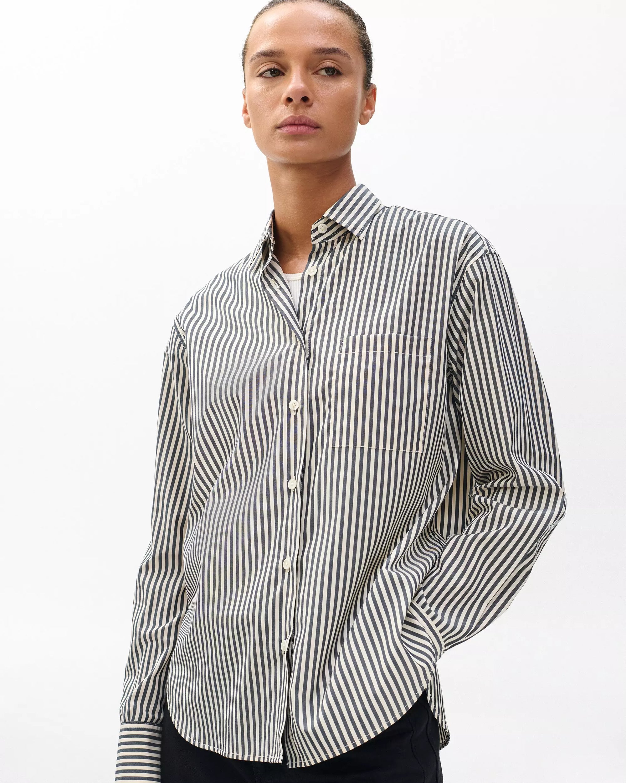 Model wearing the Rag & Bone connie striped shirt, a relaxed fit cotton poplin piece with black and white vertical stripes, stands against a plain white background with one hand in their pocket.
