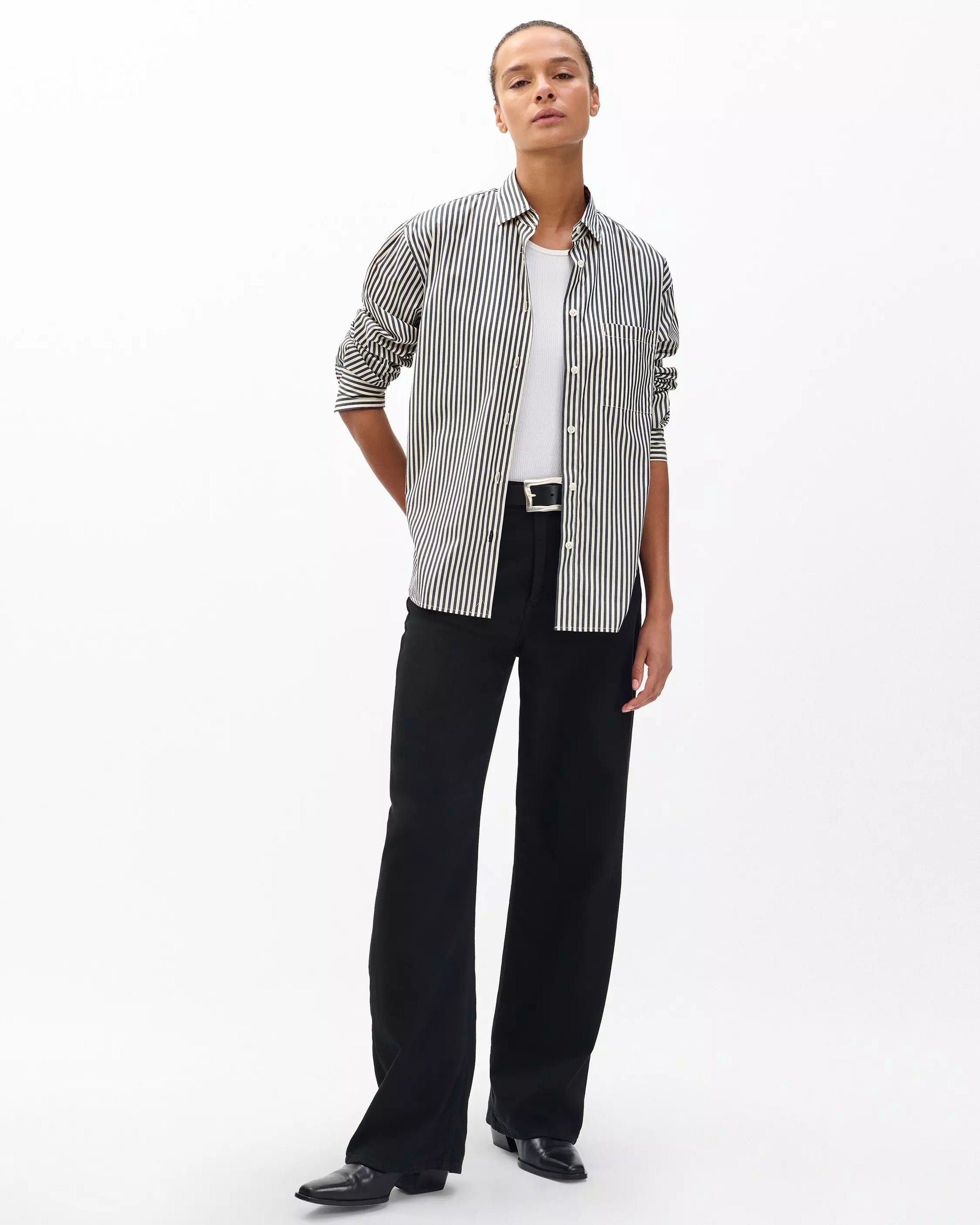 A person stands against a plain white background wearing the Rag & Bone connie striped shirt, a relaxed-fit, black-and-white cotton poplin piece layered over a white top, with black wide-leg pants and shoes. Their hands rest behind their back.