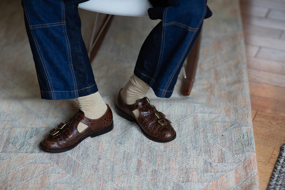 A person sits on a chair, wearing dark blue jeans and Unified Glitter Socks, paired with brown multi-buckle leather shoes, resting their feet on a patterned rug atop a wooden floor.