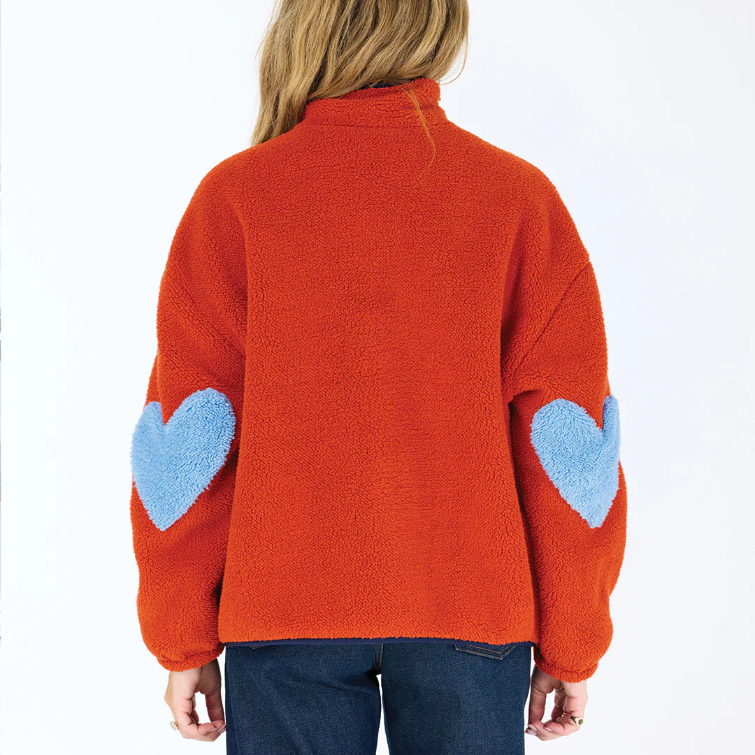 A person wearing the fuzzy orange Coze Magoze jacket by Kerri Rosenthal, featuring large light blue heart elbow patches, stands facing away from the camera against a plain white background.