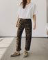 Someone stands indoors by a plain light wall, wearing a loose white tee, beige slip-on shoes, and Hey Gang’s The Officer Pant in 100% Cotton Ripstop with rolled cuffs. Their face is not visible.