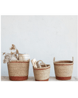 Three Creative Co-op seagrass baskets with brown stripes and handles sit against a white wall in a Scottsdale, Arizona bungalow; one contains dried flowers, another books, and the third is empty.
