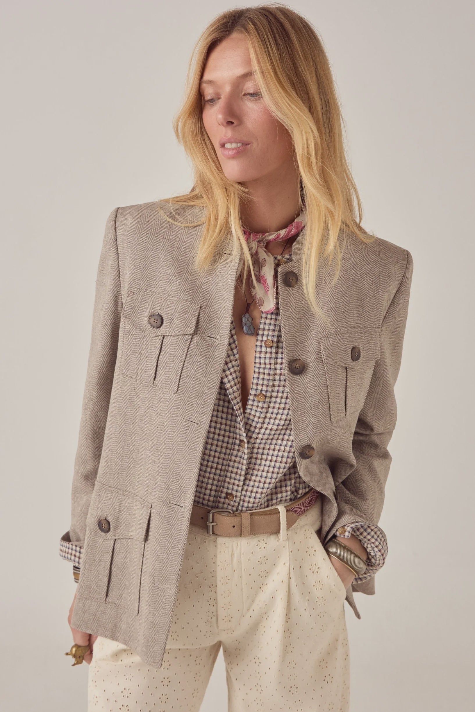 A woman with long blonde hair wears the Maison Hotel Nehru Jacket over a checked shirt, cream eyelet pants, and a patterned scarf, standing with one hand in her pocket and gazing slightly downward and to the side.