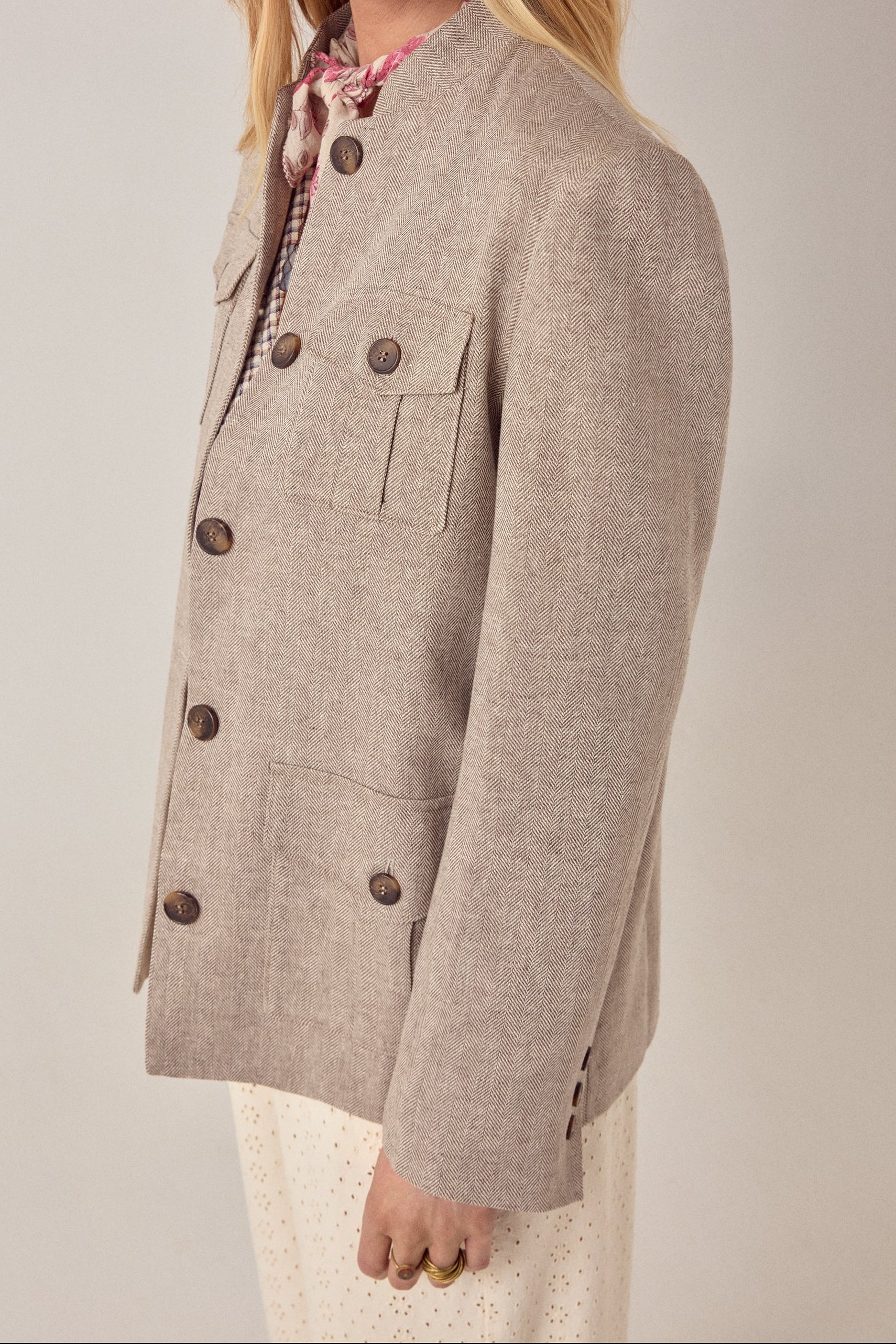 A person wearing the Maison Hotel Nehru Jacket in beige with large brown buttons, patch pockets, a printed scarf, and cream dotted pants, styled with Indian tailoring details and shown from the side against a neutral background.