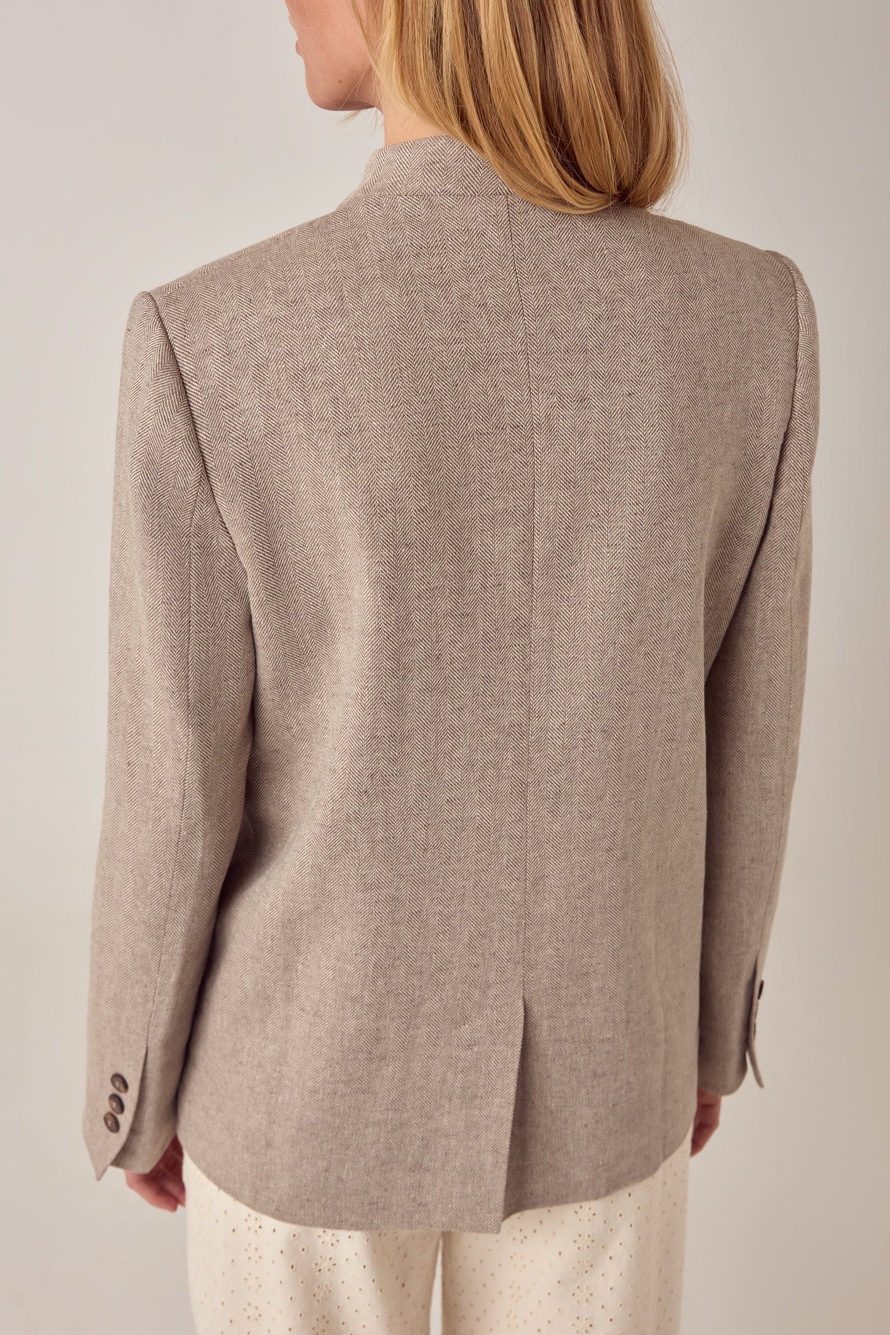 A person with long blonde hair is seen from behind wearing the Maison Hotel Nehru Jacket in light beige and perforated white pants, inspired by Indian tailoring, against a plain light-colored background.