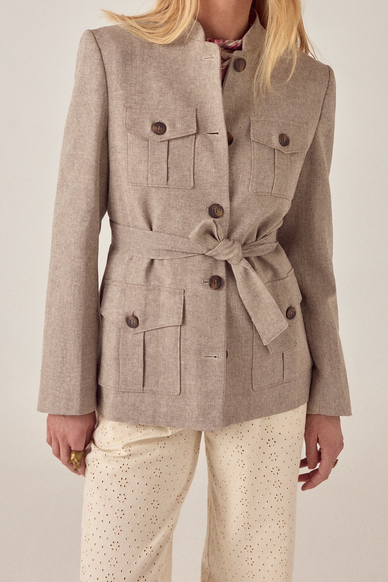A woman wears the Maison Hotel Nehru Jacket in light beige, belted and with buttoned pockets, over cream eyelet pants. Styled with loose blonde hair and relaxed hands, she stands against a neutral background.
