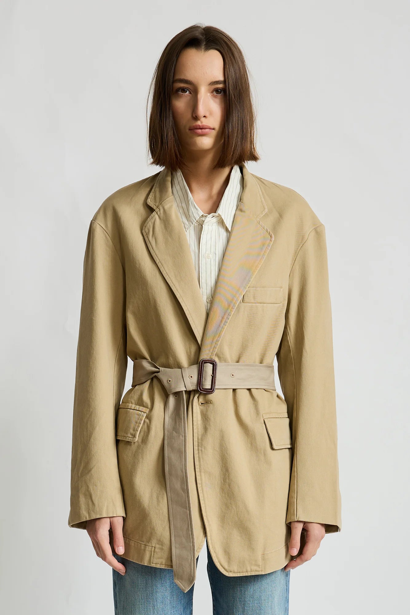 Denimist deconstructed blazer with belt in grey, styled over a striped shirt and jeans against a minimalist backdrop.