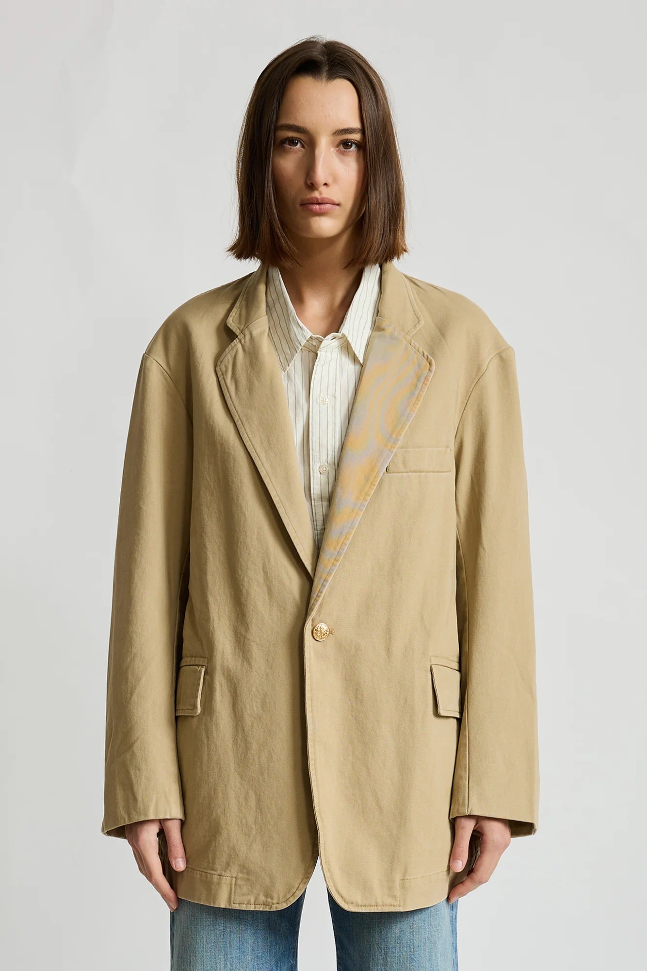 Deconstructed blazer with belt by Denimist, layered over a striped shirt and blue jeans against plain background.
