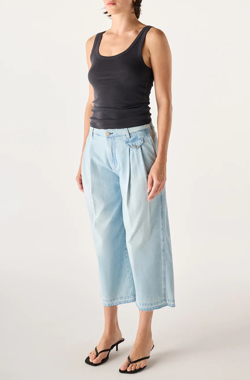 Wearing AMO Denim's Donna Trouser in light blue, a woman stands against a plain background, pairing the wide-leg crop jeans with a black tank top and black heeled sandals. Her short hair is simply styled, and her arms are relaxed at her sides.