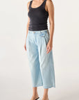 Wearing AMO Denim's Donna Trouser in light blue, a woman stands against a plain background, pairing the wide-leg crop jeans with a black tank top and black heeled sandals. Her short hair is simply styled, and her arms are relaxed at her sides.