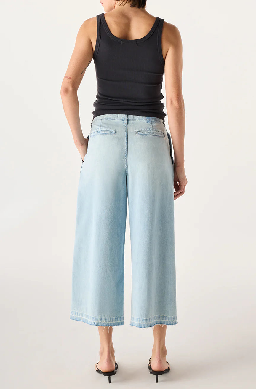 A woman stands facing away against a plain light background, wearing the AMO Denim Donna Trouser—a crop-length wide leg jean—paired with a black sleeveless tank top and black heeled sandals.