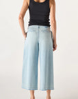 A woman stands facing away against a plain light background, wearing the AMO Denim Donna Trouser—a crop-length wide leg jean—paired with a black sleeveless tank top and black heeled sandals.
