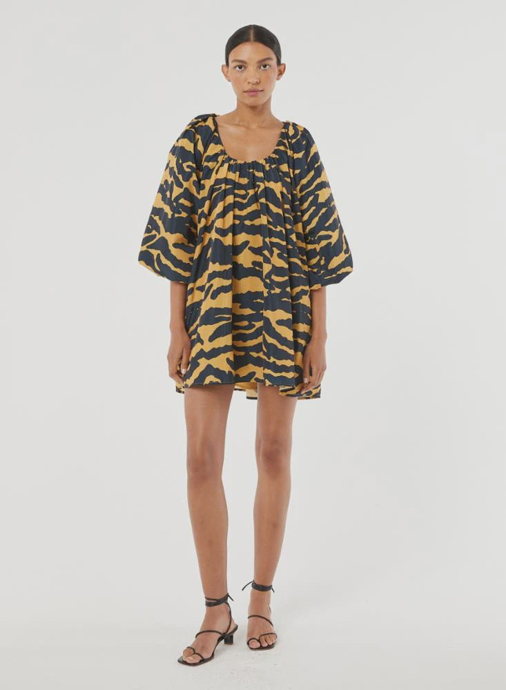 A person stands wearing the loose-fitting Alina Dress by Rhode, featuring an abstract print, bubble half-sleeves, and a gathered neckline. Crafted from recycled polyester, the dress is paired with black sandals and a neutral expression against a plain white background.