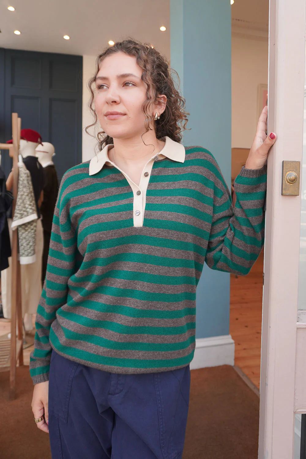 A woman with curly hair stands indoors, wearing the Jumper 1 2 3 4/CR2 Merino Rugby Shirt—an oversize top with green and gray stripes, a light collar—and blue pants. She rests one hand on a door frame, gazing thoughtfully into the distance.