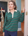 A woman with curly hair stands indoors, wearing the Jumper 1 2 3 4/CR2 Merino Rugby Shirt—an oversize top with green and gray stripes, a light collar—and blue pants. She rests one hand on a door frame, gazing thoughtfully into the distance.