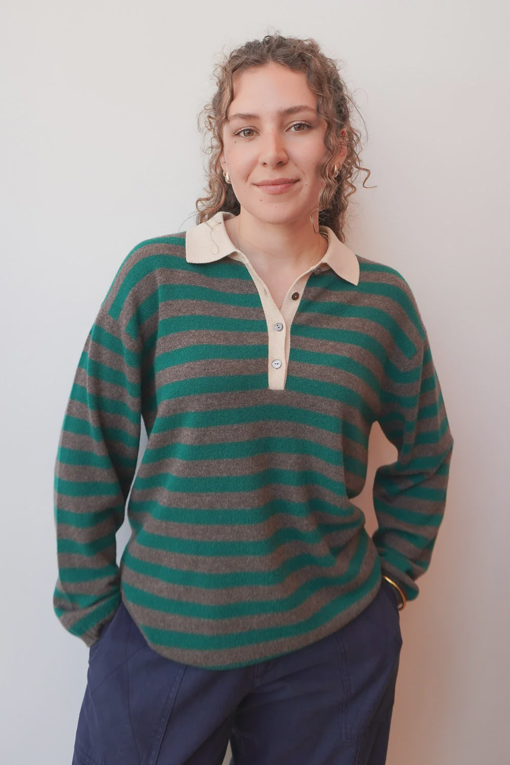 A woman with curly hair wears the Jumper 1 2 3 4/CR2 Merino Rugby Shirt in green and brown stripes, made of soft merino wool. She pairs it with dark pants and stands against a light wall, smiling softly with her hands in her pockets.
