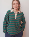 A woman with curly hair wears the Jumper 1 2 3 4/CR2 Merino Rugby Shirt in green and brown stripes, made of soft merino wool. She pairs it with dark pants and stands against a light wall, smiling softly with her hands in her pockets.