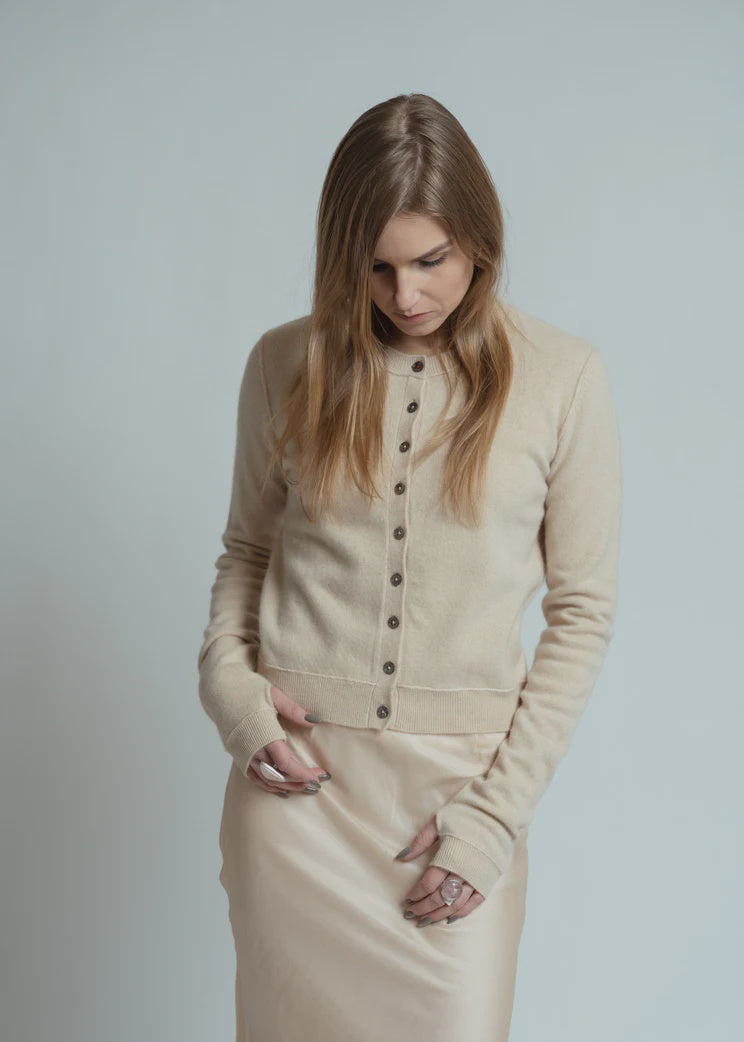 A woman with long blonde hair wears the Brazeau Tricot Royal All Thumbs Cardigan in beige, featuring a button front, paired with a light beige skirt. She stands hands in pockets against a plain light background, looking down.